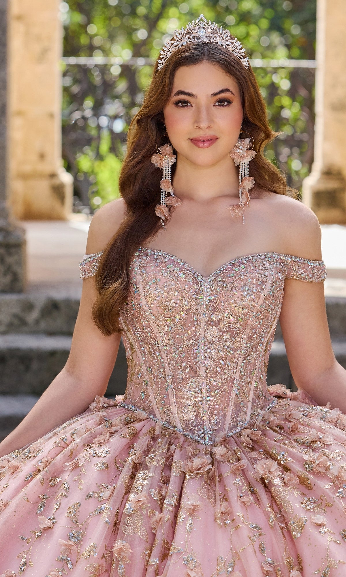 Wearing the Princesa PR30131 off-shoulder quinceanera dress adorned with sequins and beading, a young woman in a tiara and long earrings poses outdoors in formal attire.